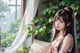 A woman with long hair and traditional attire sits by a window with greenery.